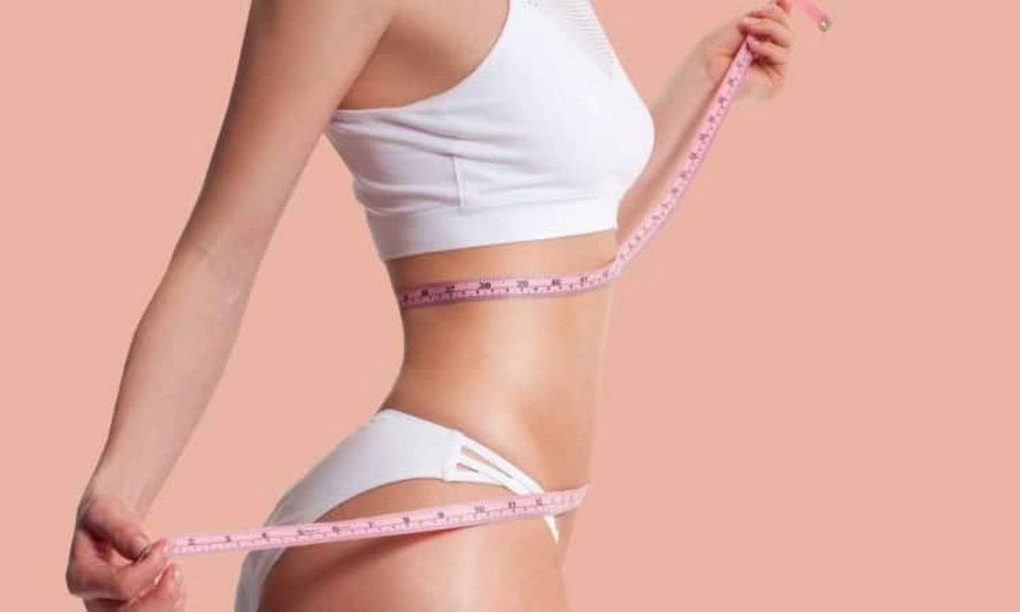 Woman measuring her waist and hips with a pink measuring tape, showcasing a slim body shape in white activewear.