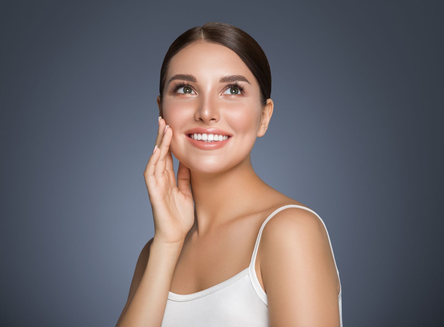 Woman with glowing skin smiling and touching her cheek against a dark background