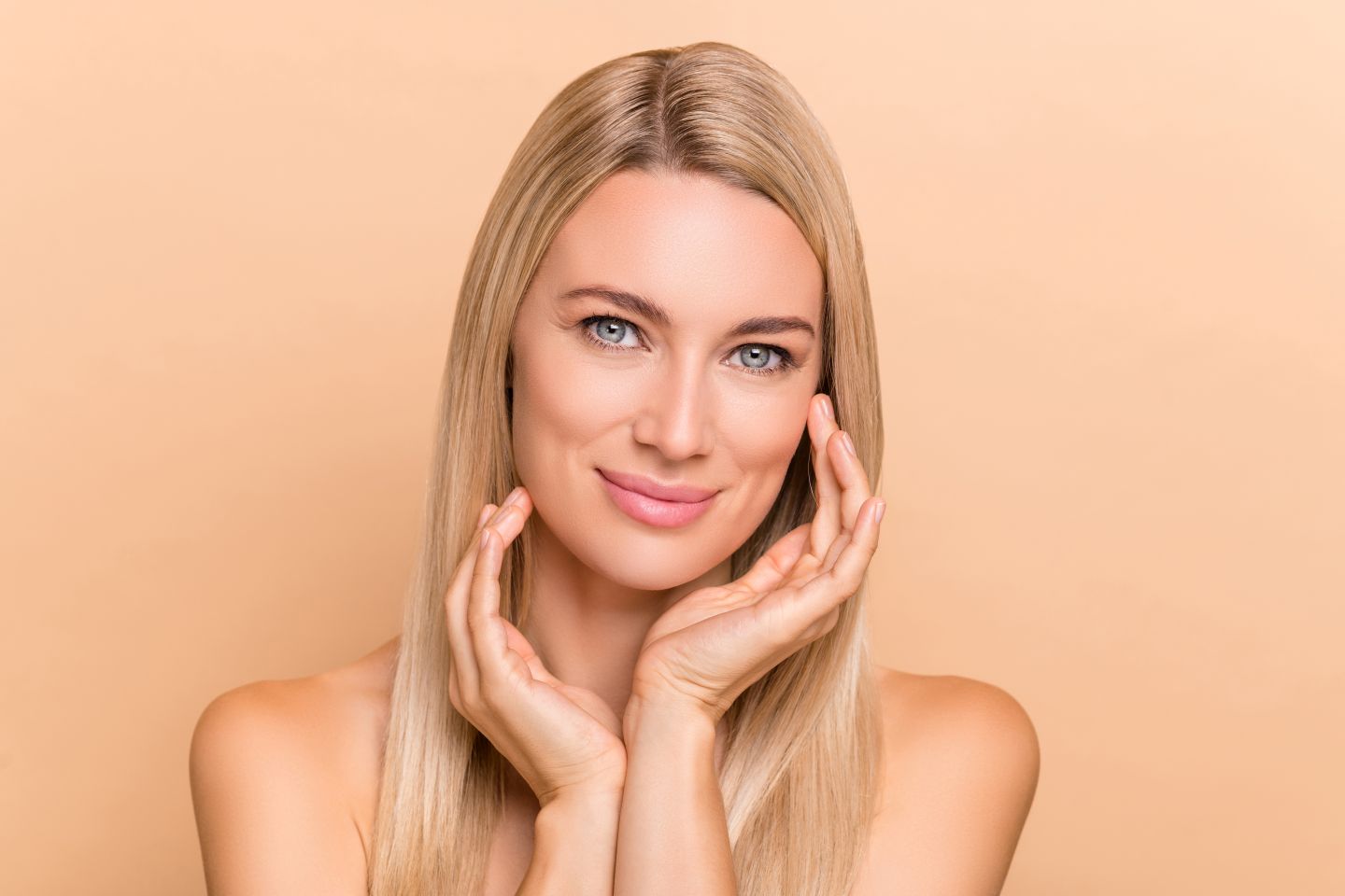 Smiling blonde woman with smooth skin touching her face against a peach background.